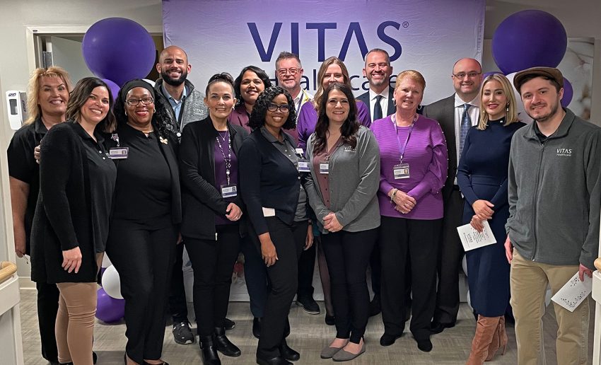 A group of people stands in front of a VITAS sign in the inpatient unit, with purple balloons